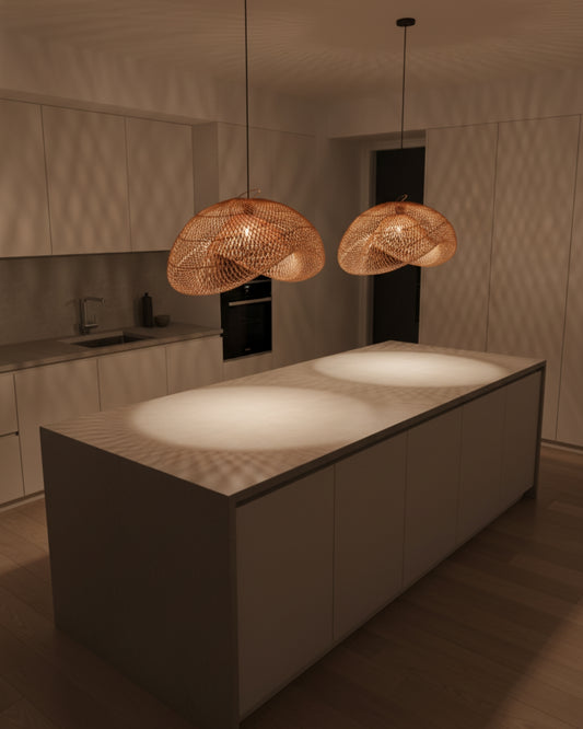 Two handwoven rattan pendants with sculptural wave shades over a kitchen island at night; 3000K warm glow creates patterned pools on the countertop and walls. Black canopies and cords in a minimal Japandi kitchen.