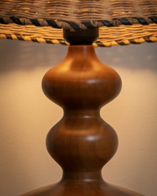 Close-up of sculpted ash-wood lamp base in walnut tone beneath a handwoven rattan shade with braided edge; smooth satin finish and warm 2700K glow highlighting wood grain and weave texture on a bedside table.