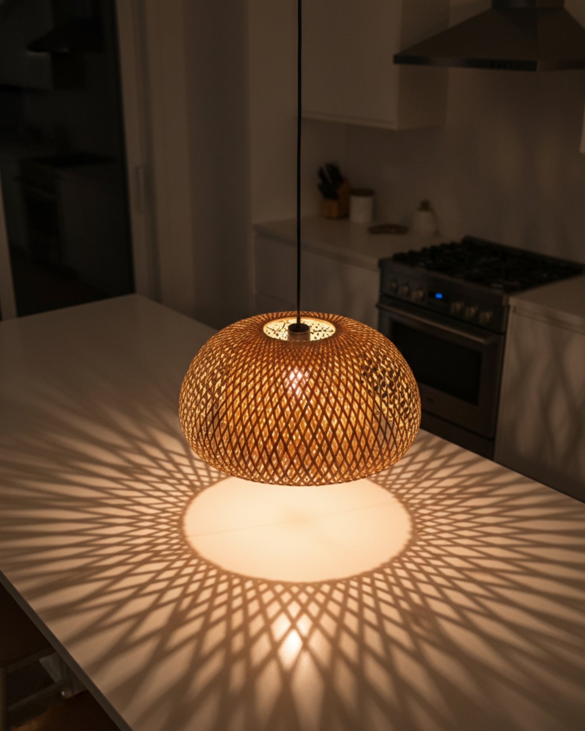 Handwoven bamboo dome pendant above a kitchen island, lamp on at 2700K. Top-down view with a crisp radial lattice shadow on the white countertop; straight black cord, modern kitchen background.
