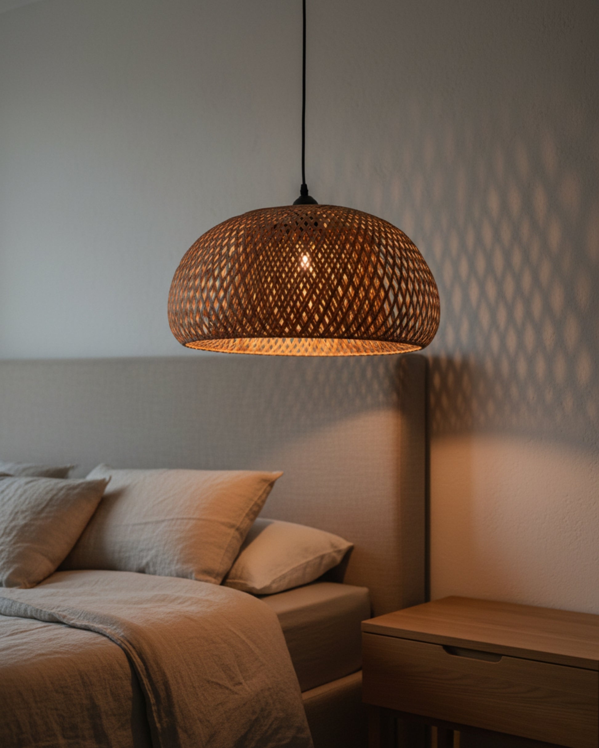 Handwoven bamboo dome pendant beside a bed, lamp on at 2700K; straight black cord and canopy. Soft lattice shadows on the wall, warm glow over linen bedding and oak nightstand in a calm Japandi bedroom.