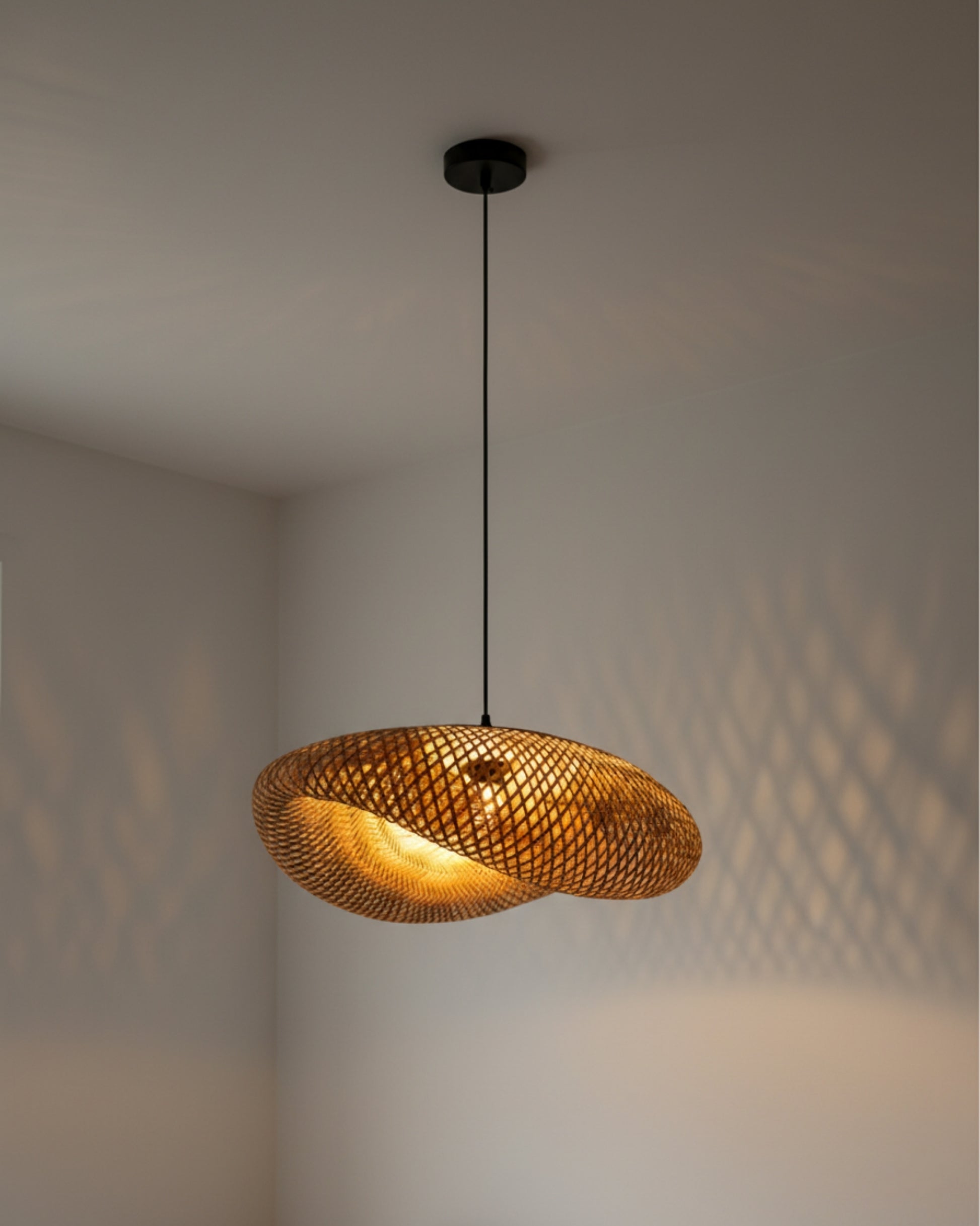Evening view of a handwoven bamboo twisted-disc pendant, ~31.5 in (80 cm) diameter with black canopy and slim cord; warm 2700K bulb ON casting lattice shadows on the wall; ideal above a kitchen island or dining table in Japandi interiors.