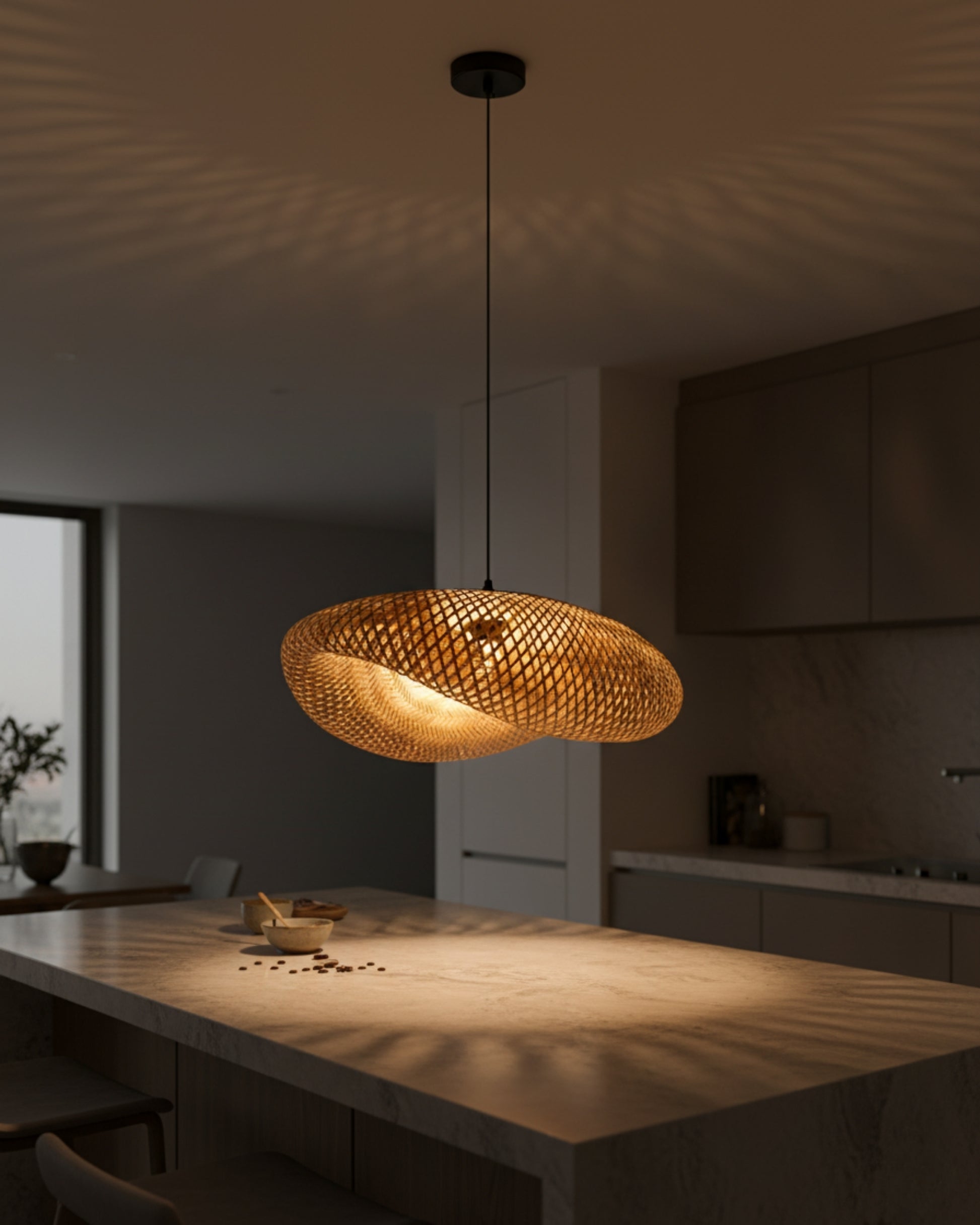Kitchen island evening view of a handwoven bamboo twisted-disc pendant, approx. 31.5 in (80 cm) with black canopy and slim cord; warm 2700K bulb ON casting lattice shadows across the countertop; Japandi/Scandinavian interior.