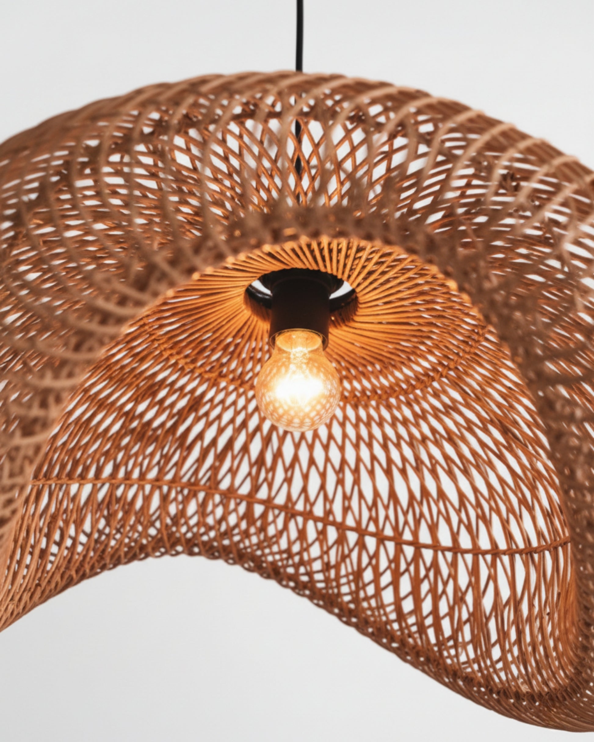 Handwoven rattan pendant with a sculptural wave shade, close-up looking up at the glowing 3000K bulb and black socket; airy woven lattice casts patterned light—perfect above a kitchen island or dining table.