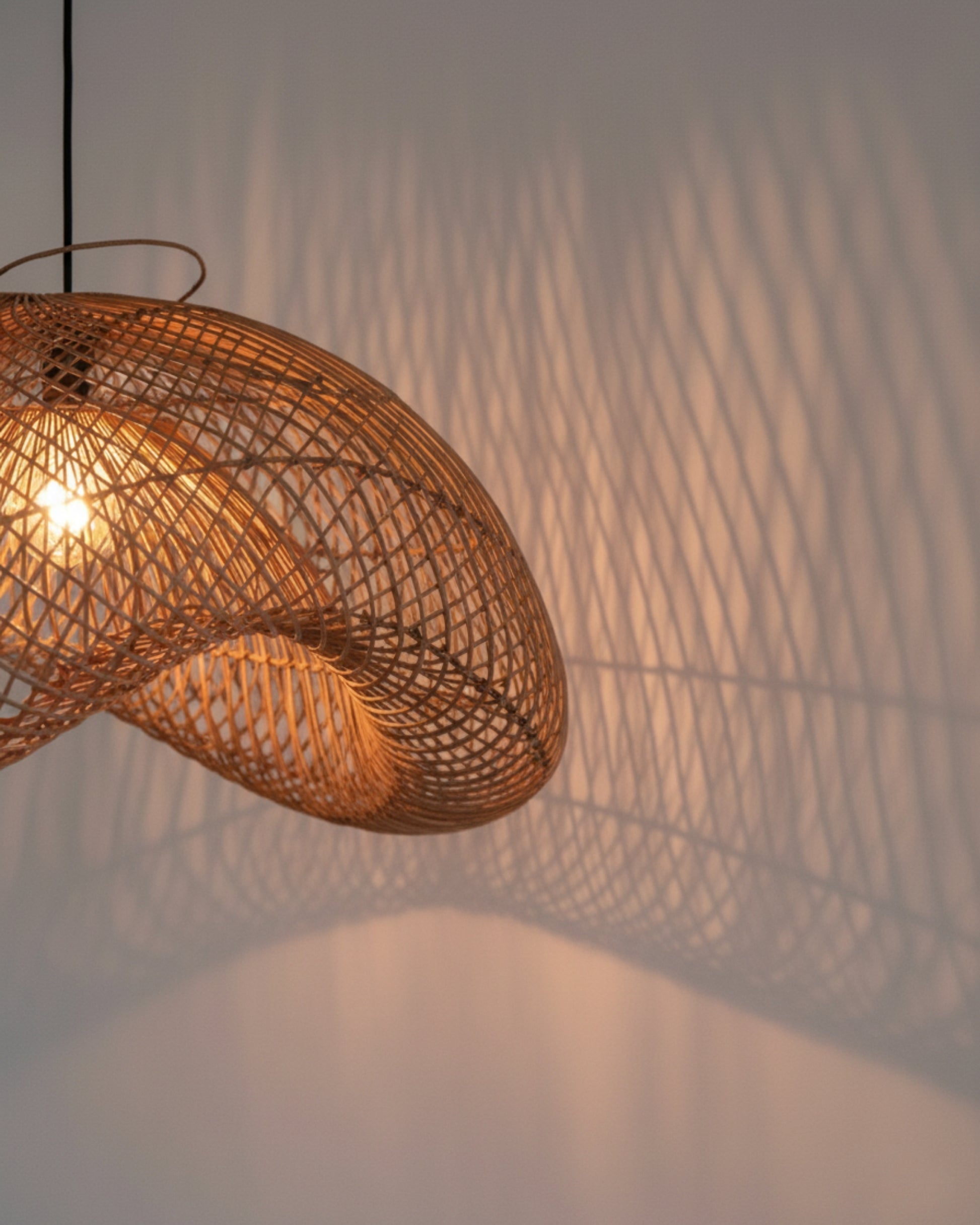 Handwoven rattan pendant with sculptural wave shade, warm 3000K bulb on—casting patterned lattice shadows on the wall; natural honey-brown weave and airy texture, perfect for ambient lighting in Japandi living rooms, bedrooms, or dining nooks.