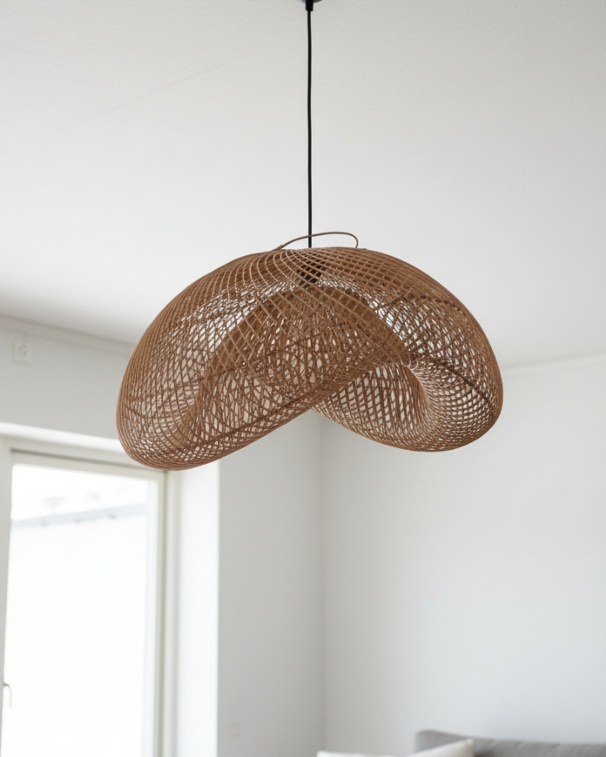 Daylight view of a handwoven rattan pendant with a sculptural wave shade hanging in a bright living room; natural honey-brown weave with black cord and canopy, airy lattice that casts soft patterns—ideal for Japandi/Scandinavian interiors.