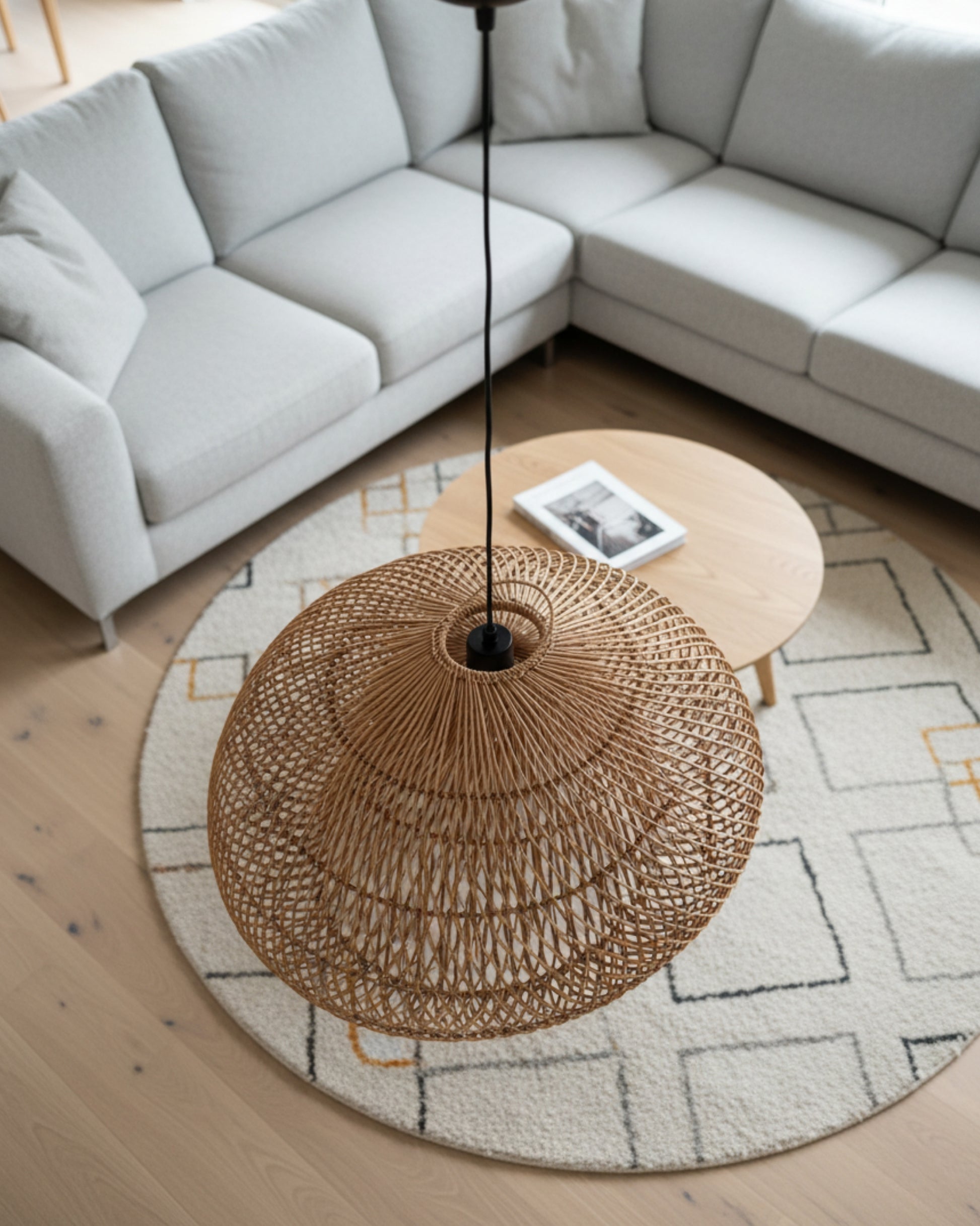 Top-down view of a handwoven rattan pendant with spiral lattice and black cord, hanging over a Japandi living room—round textured rug, light gray sectional, and oak coffee table; lamp off in soft daylight, airy weave adds warm texture.