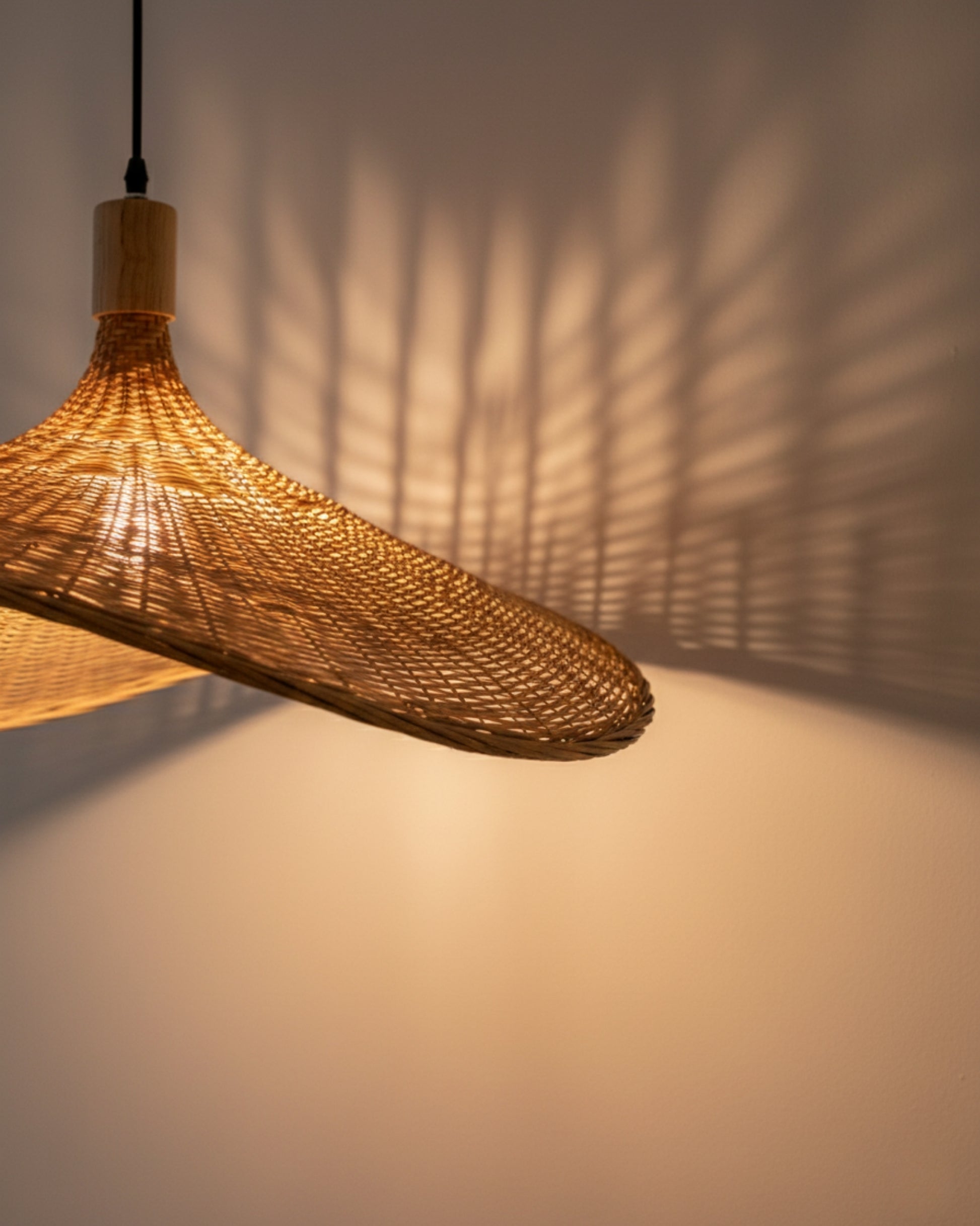 Handwoven rattan pendant light with a sculptural wave shade, lamp on at 2700K; open weave throws a dramatic lattice shadow across the wall—warm, organic ambiance for a kitchen island or dining table.