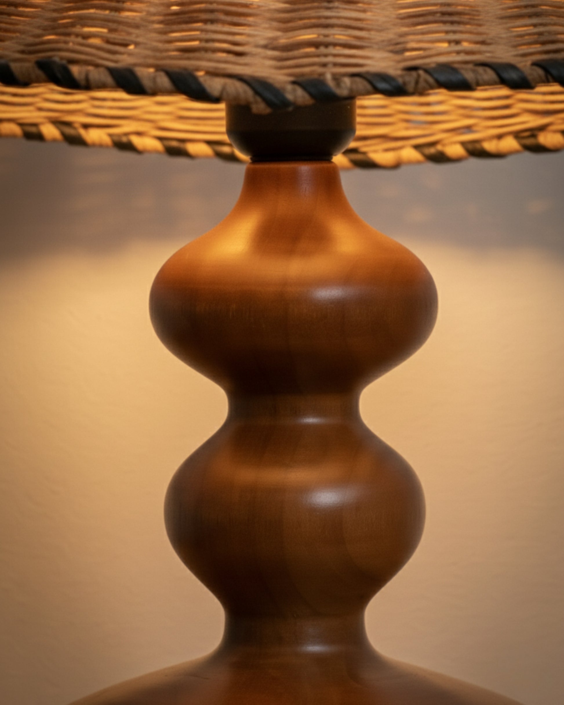 Close-up of sculpted ash-wood lamp base in walnut tone beneath a handwoven rattan shade with braided edge; smooth satin finish and warm 2700K glow highlighting wood grain and weave texture on a bedside table.