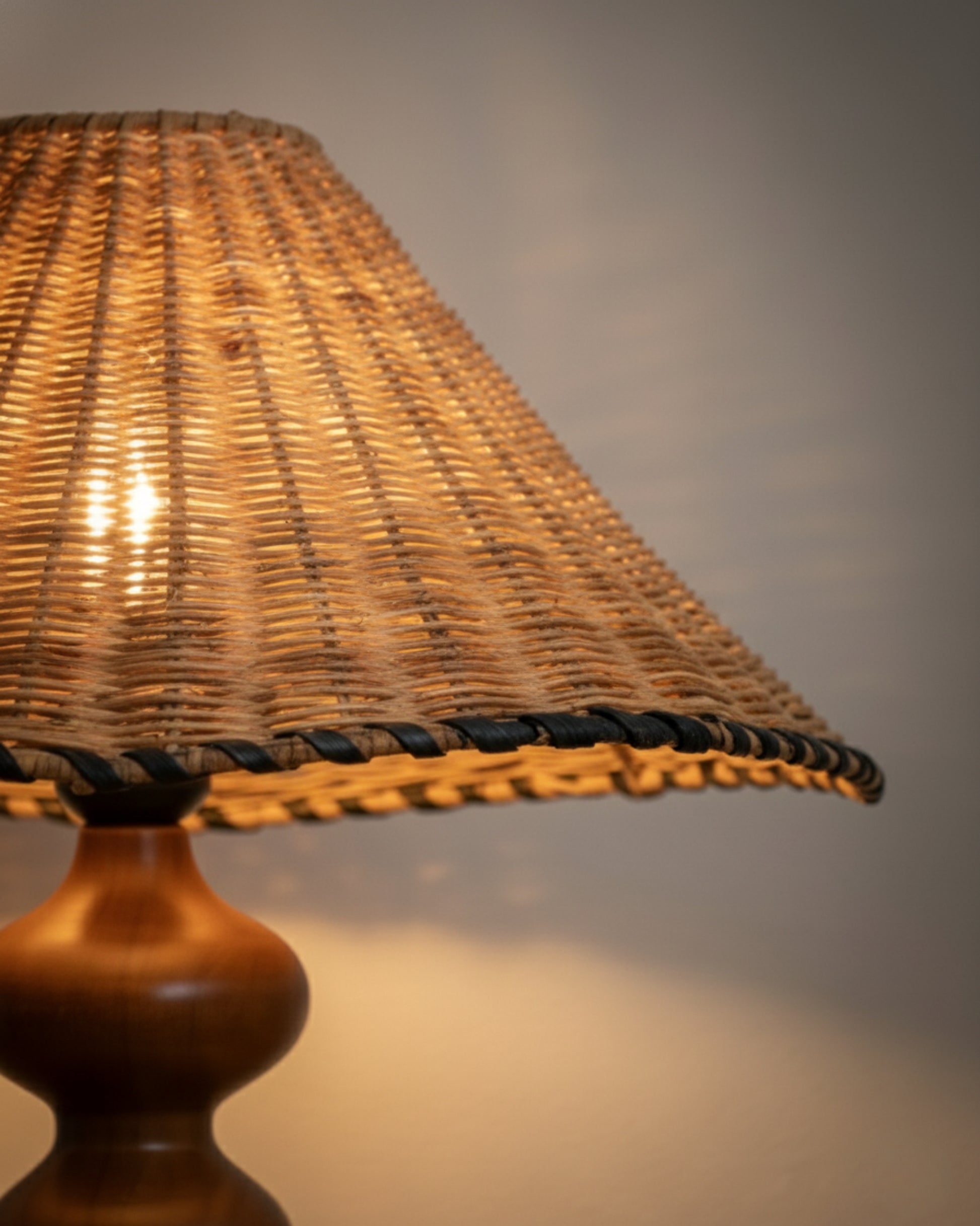 Angled view of the handwoven rattan lampshade with braided black edge; warm 2700K light shines through the weave. Paired with a sculpted ash-wood base in walnut tone; 40 cm (15.75") shade casting a soft ambient glow on the wall.