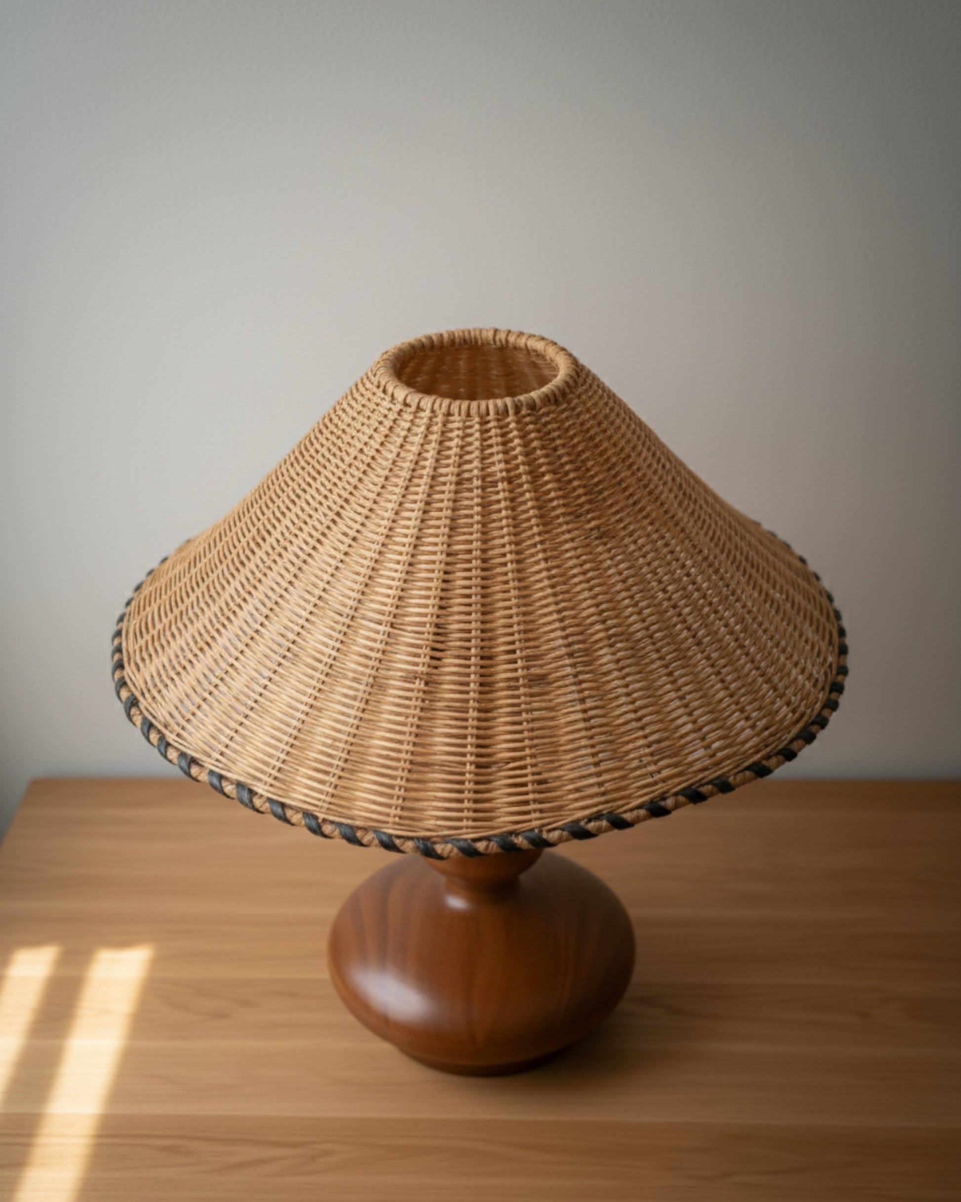 Top-down view of a handwoven rattan table lamp with sculpted ash-wood base (walnut tone); 400 mm (15.75") conical shade with braided edge on a light-wood tabletop in natural daylight; ideal for Japandi bedrooms or living rooms.