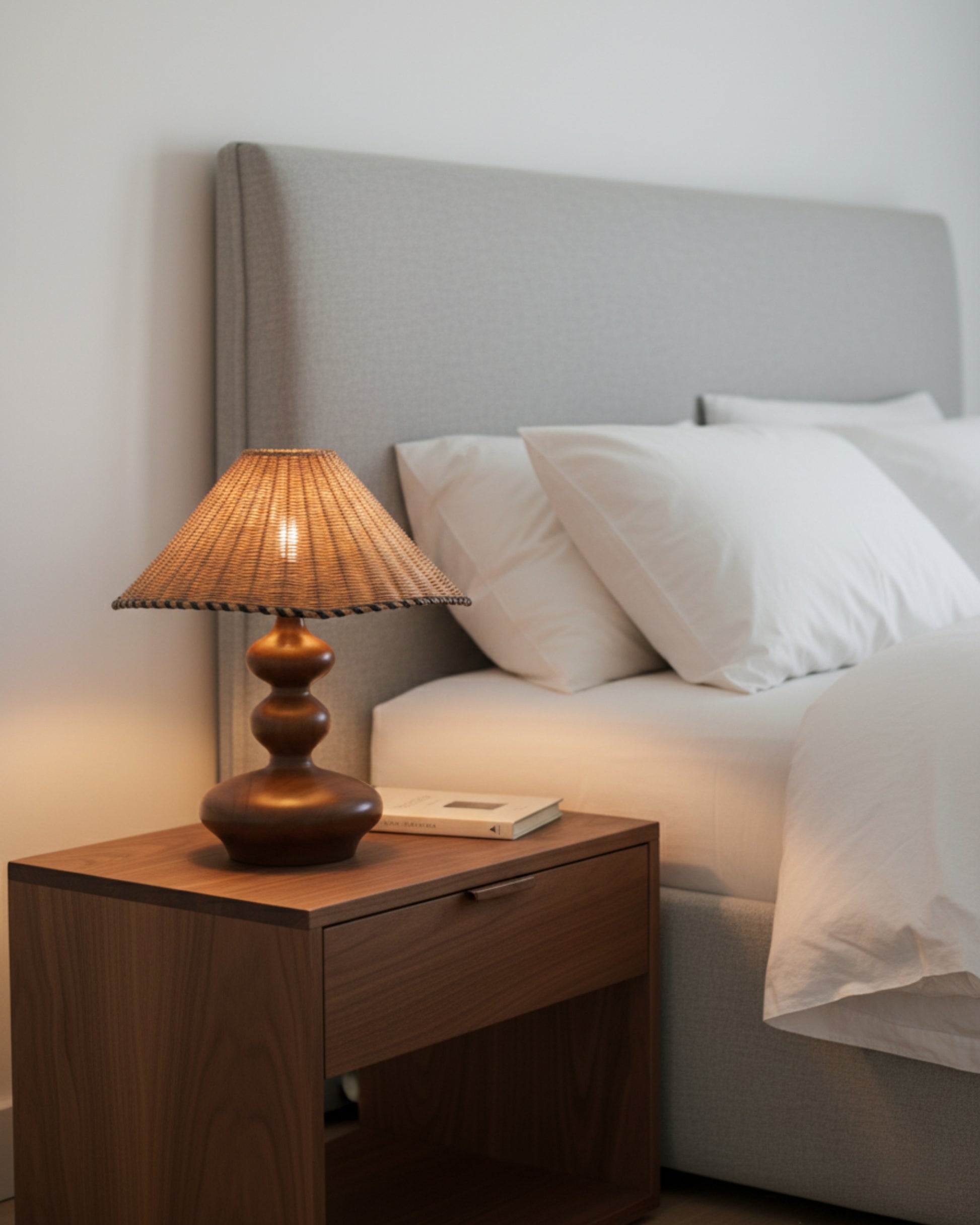 Handwoven rattan table lamp with sculpted ash-wood base (walnut tone), 40 cm (15.75") shade, lit on a walnut nightstand beside a grey upholstered bed; soft 2700K ambient glow for Japandi bedrooms.