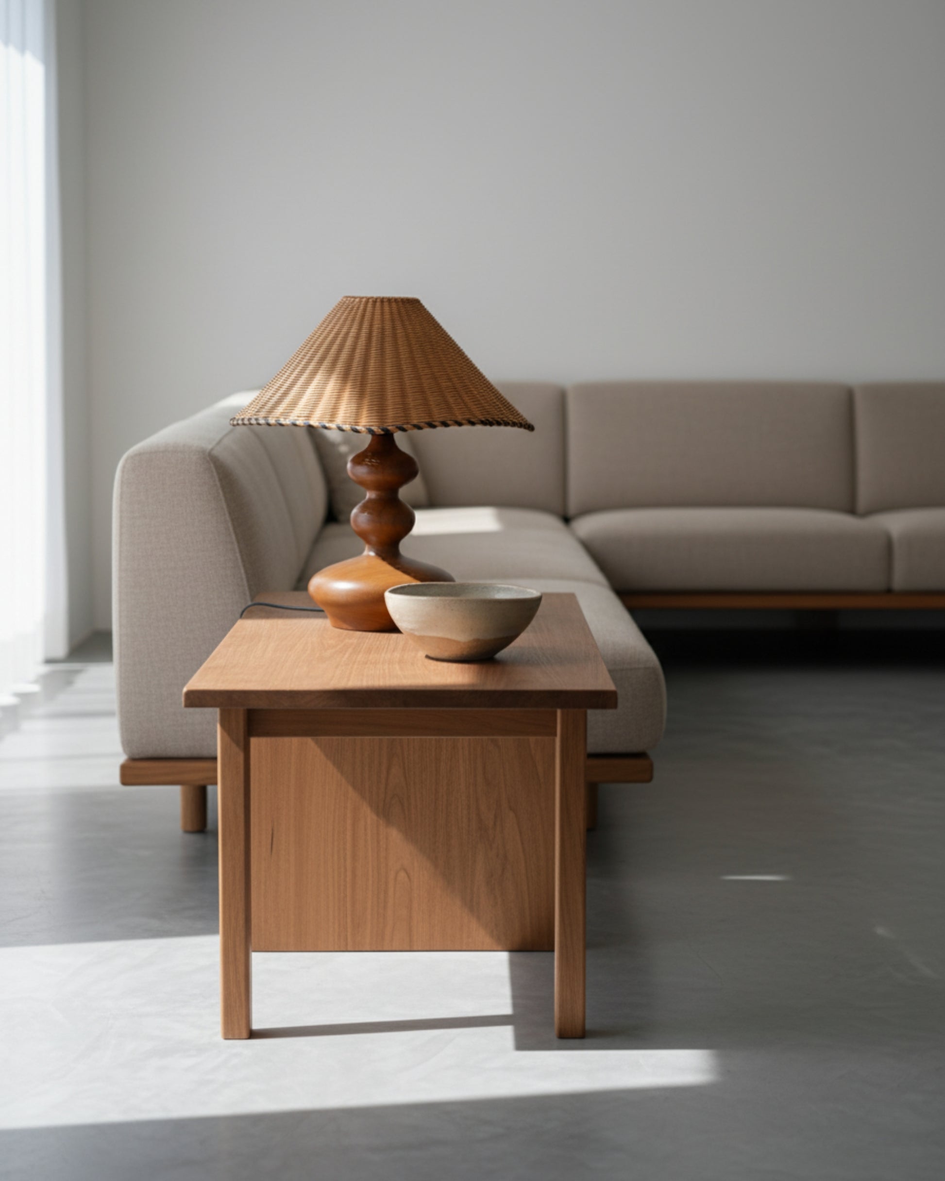 Handwoven rattan table lamp with sculpted ash-wood base (walnut tone), 40 cm (15.75") shade, on a low wood side table next to a beige sectional sofa; soft daylight in a minimalist Japandi living room.